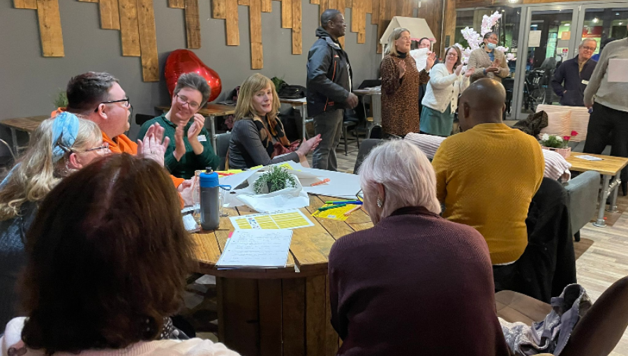 Inside a cafe, groups of people at round tables sit clapping as several speakers stand at the front of the room talking about their ideas for community led change in Coventry. This is Collaboration Station.