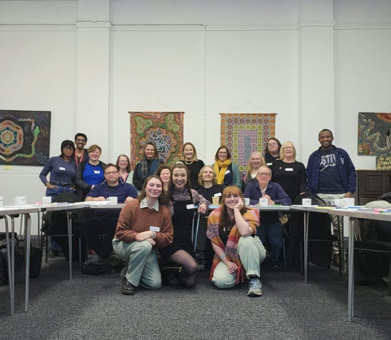 A large group of people gather around a table in a community space in Coventry for a photo during community organising training. Some are seated, some standing and some kneeling at the front for the group shot.