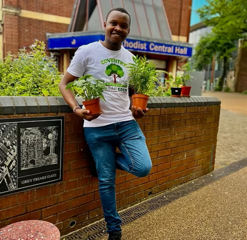 A young black man with short hair and a clean shaven face smiles as he holds potted plants next to a raised flower bed in Coventry city centre. He is Ade - a member of Coventry Urban Eden, whose branded white t-shirt he is wearing with blue jeans.