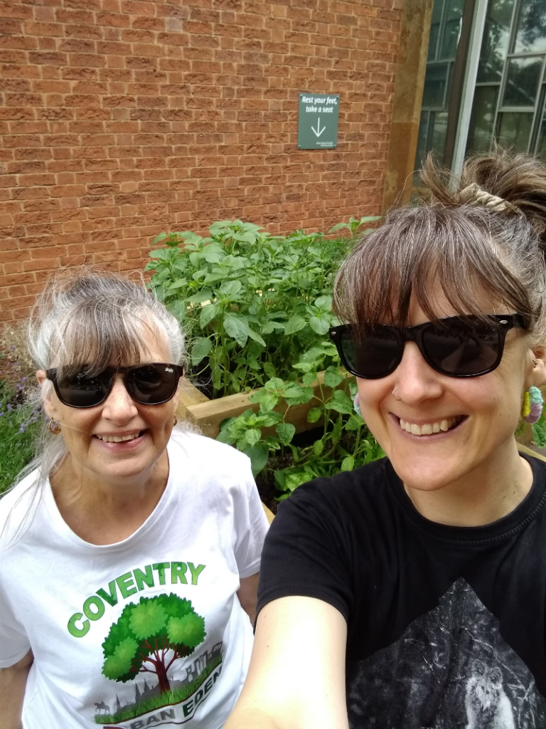 two white women in sunglasses smiling for a selfie in front of newly planted wooden planters outside the Herbert Art Gallery & Museum in Coventry. One woman wears a white Coventry Urban Eden t-shirt with green tree logo.