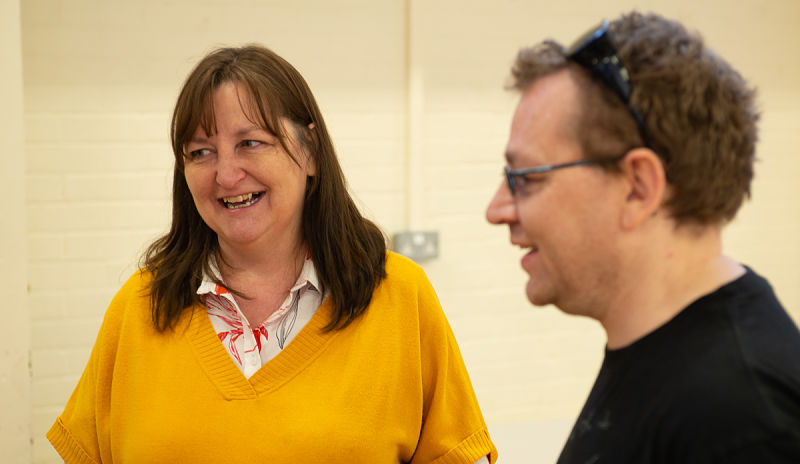 A woman with brown hair and wearing a yellow jumper talks to a man in glasses indoors in Willenhall, Coventry