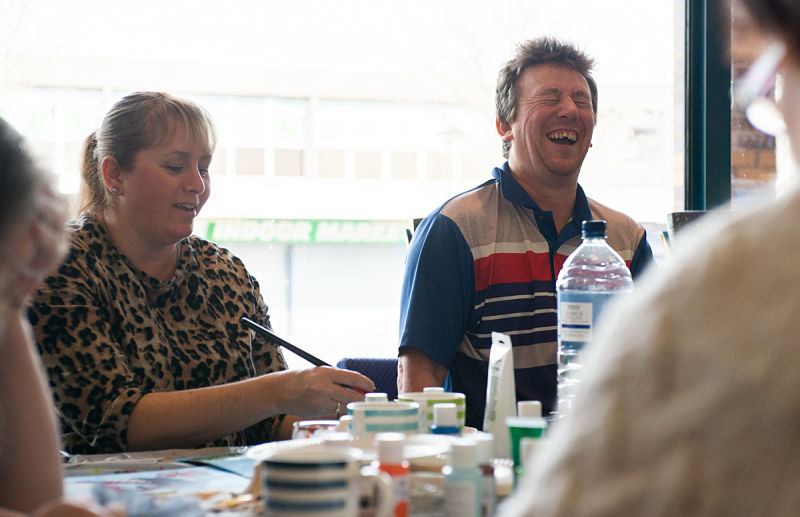 A woman and a man sit together painting in a library setting. The man is laughing out loud with his eyes closed