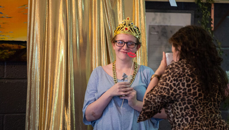 A young woman poses for the camera wearing a dress up crown in front of a gold curtain.