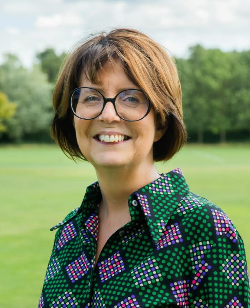 A smiling older white woman with bobbed brown hair and large round black rimmed glasses wears a patterned pink and green shirt for a photo outdoors among grass and trees.