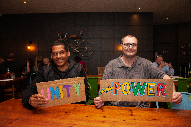 Two men, one white and one Asian, sit side by side at a coffee table holding signs saying Unity and Power.