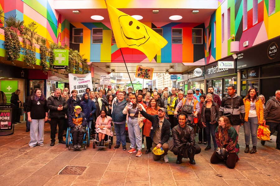 A large group of around 50 diverse people stand below a rainbow-coloured roof and a giant yellow smiley face flag in a Coventry city centre shopping precinct. Megaphones and banners can be also be seen.