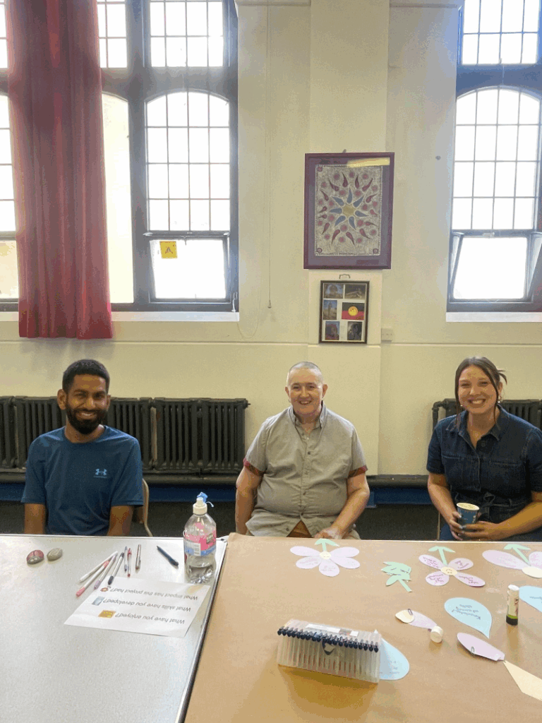 Three people smile as they sit at a table in a hall with craft materials before them. From left to right, they are experts by experience Huzaifah and Alix with Grapevine project worker Sophie.