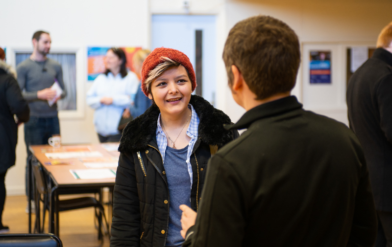 A young woman wearing a red beanie hat smiles as she talks to a man with his back to the camera. In the background is a community centre and other people talking.