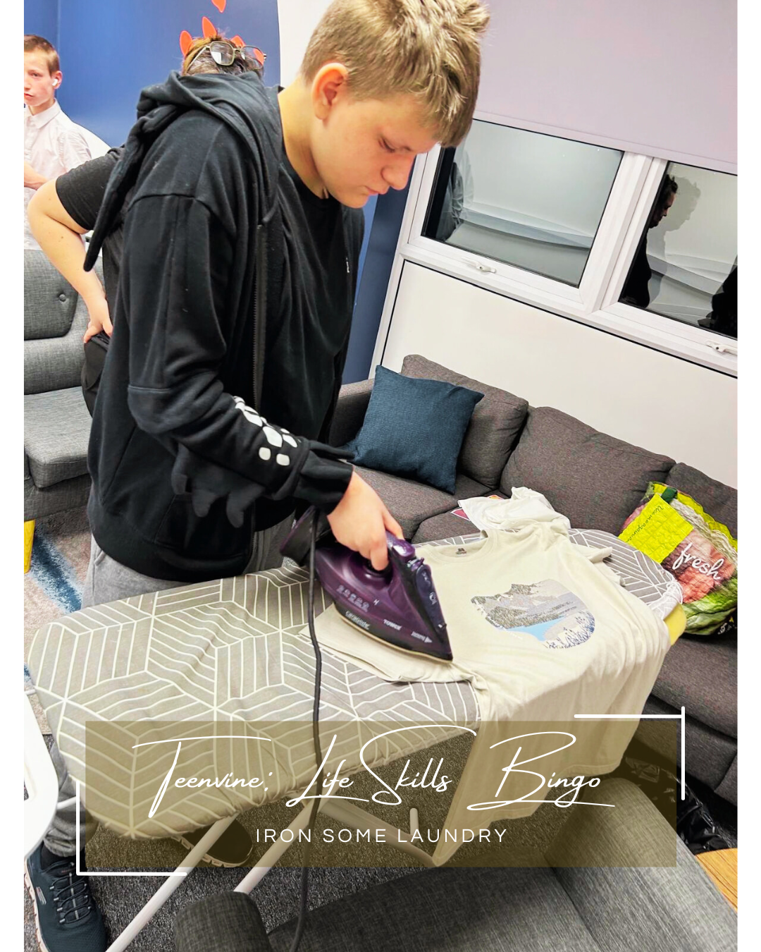 A young white disabled boy wears a black hoodie and uses an iron and ironing board to iron some clean clothes in a community venue.