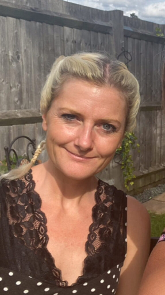 A blonde middle-aged woman with plaits and wearing a black vest smiles for a photo in front of a garden fence. The sun is shining.