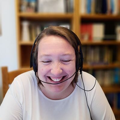 A smiling white woman wearing headphones with microphone works from home during the pandemic. A blurred bookcase is seen in the background.