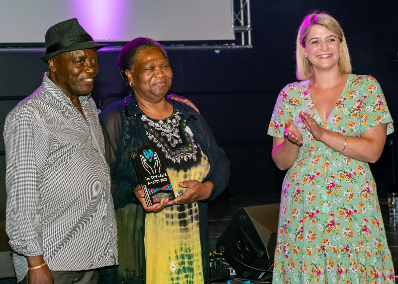 An older black couple stand smiling while holding their Cov Cares award for best venue for the Knights Bar at Two Tone Village. A white woman wearing a flowery dress with bobbed blonde hair claps after presenting them with the award.