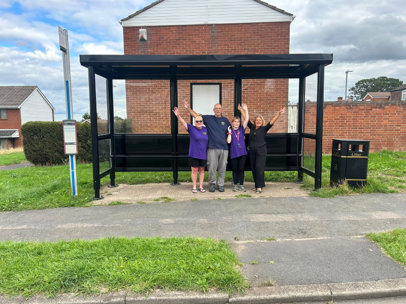 Three white women and one white man raise their arms in celebration under a new bus shelter in Nuneaton. They are some of the Friends of Grove Farm (left to right: Gwyn, Paul, Vicky and Grapevine community organiser Michelle).