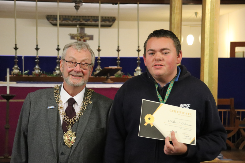 Nathan wears a black jumper and holds his award certificate as he stands next to the Lord Mayor, an older man with glasses, grey hair and beard and gold chains of office.