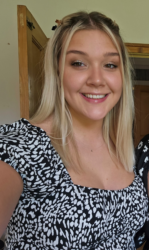 A white woman in her twenties with shoulder-length blonde hair and wearing a black and white print top, smiles for the camera with an open wooden door behind her. She is Lucy from Grapevine.