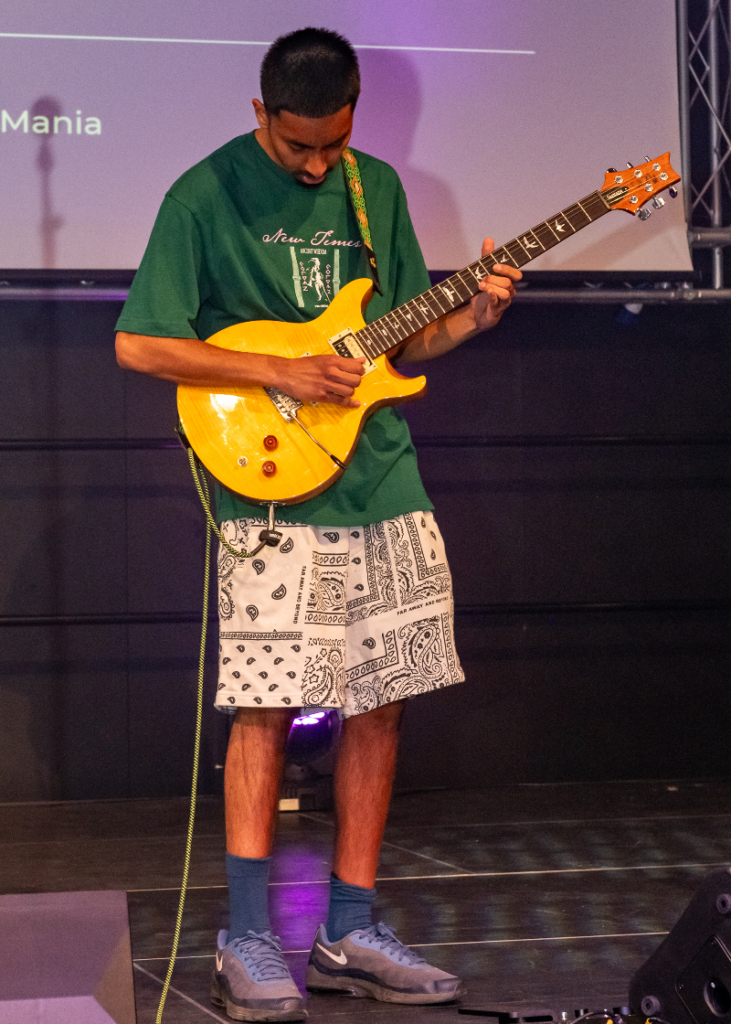 A young Asian man wears shorts and a green T-shirt and plays a yellow electric guitar on stage at the event.