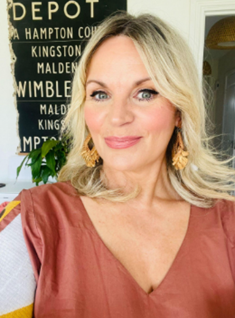 A white woman in her fifties with long blonde hair and wearing an orange sun dress and gold earrings smiles for a selfie in her home.