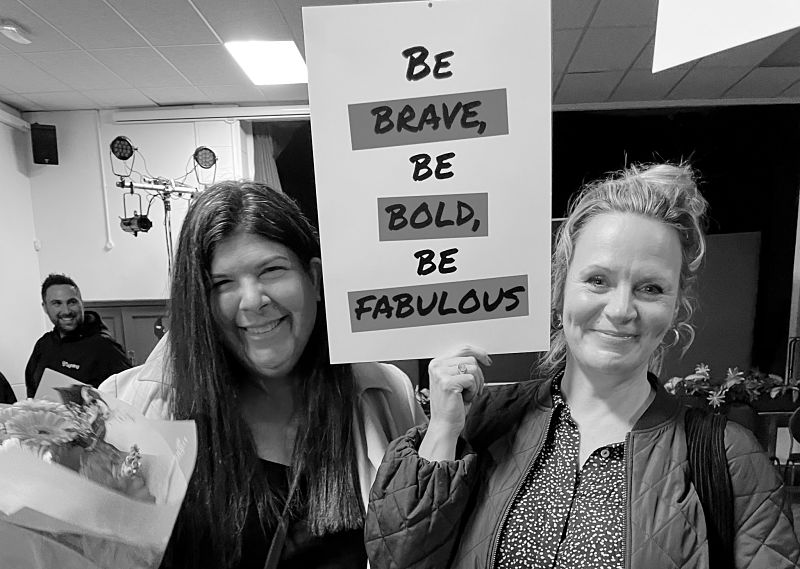 A black and white photo of two women, one blonde and the other with long dark hair, holding a sign that says Be brave, Be bold, Be fabulous