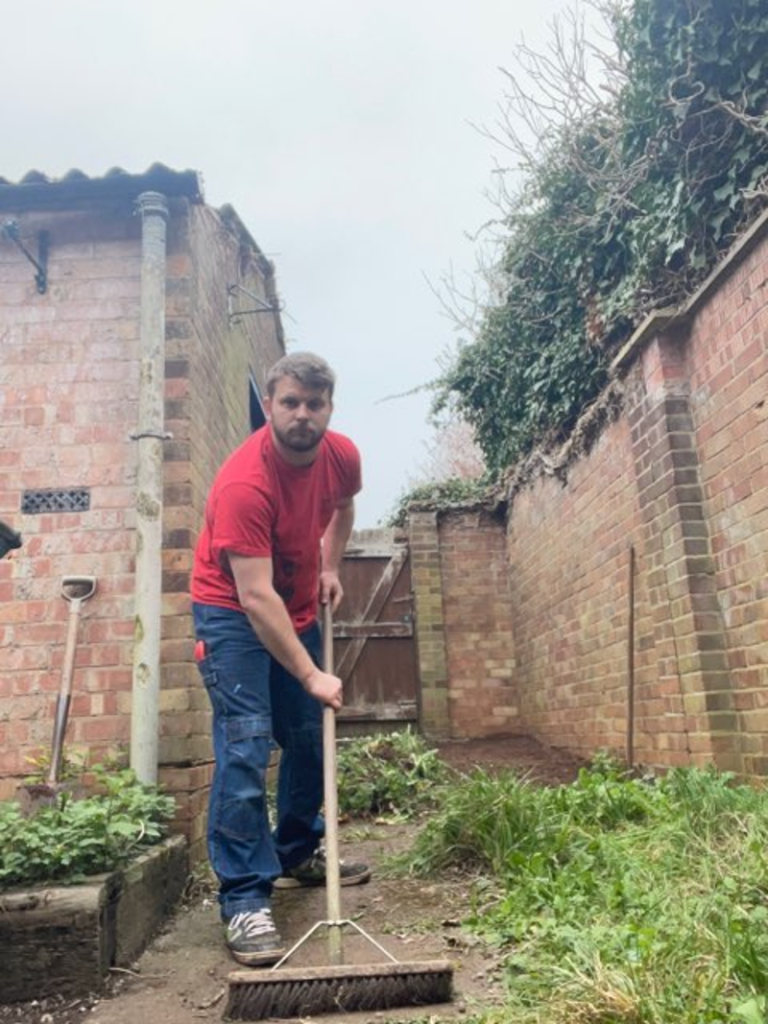 Michael wears a red t-shirt and holds a wooden brrom to sweep the backyard of a house in Allesley village, Coventry. Brick walls and outbuildings can be seen.