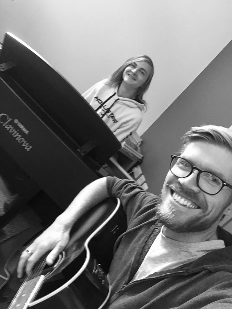 A man in glasses plays guitar with a young man on piano