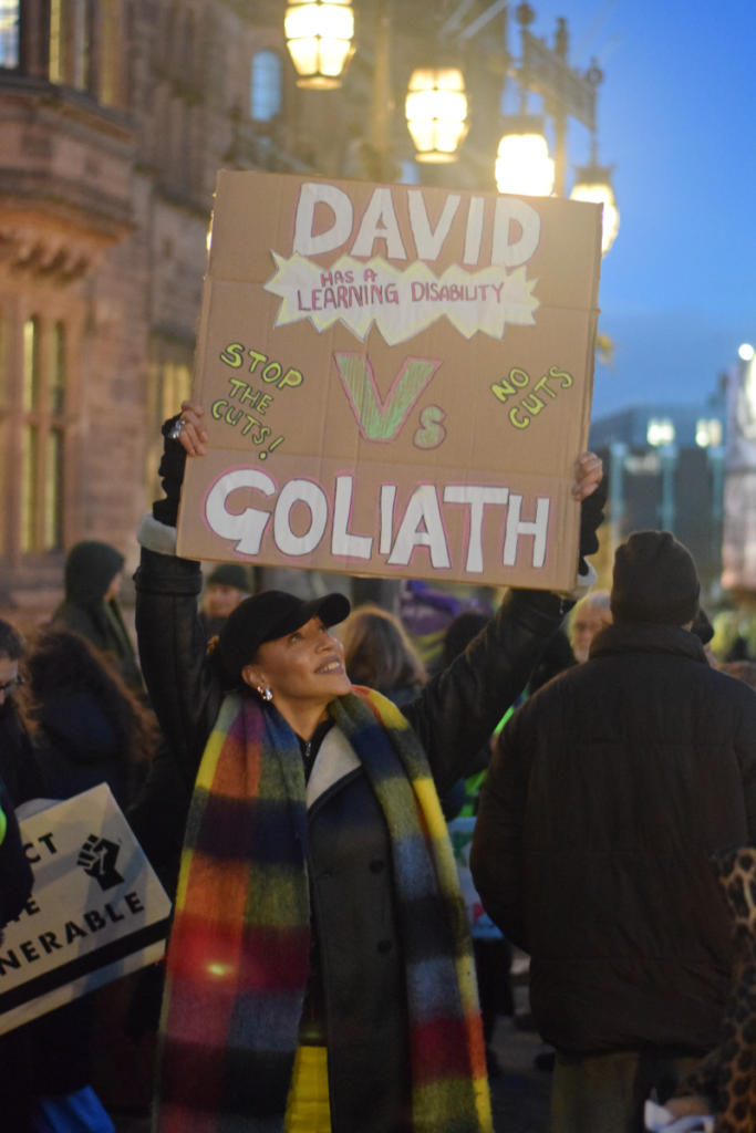 Help and Connect lead and our Director of Projects, Naomi Madden, wears a brightly checked scarf with black baseball cap as she holds a handmade cardboard placard reading, "David (has a learning disability) versus Goliath. Stop the cuts!"