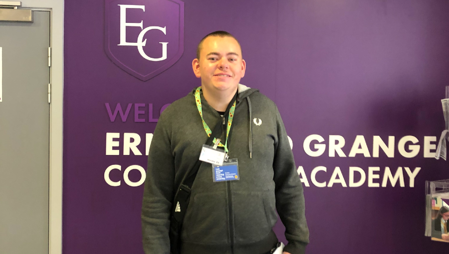 Nathan, a young man with closely cut brown hair wears two lanyards and a crossbody bag as he stands in front of a purple Ernesford Grange sign inside the school. He is smiling on his first day.