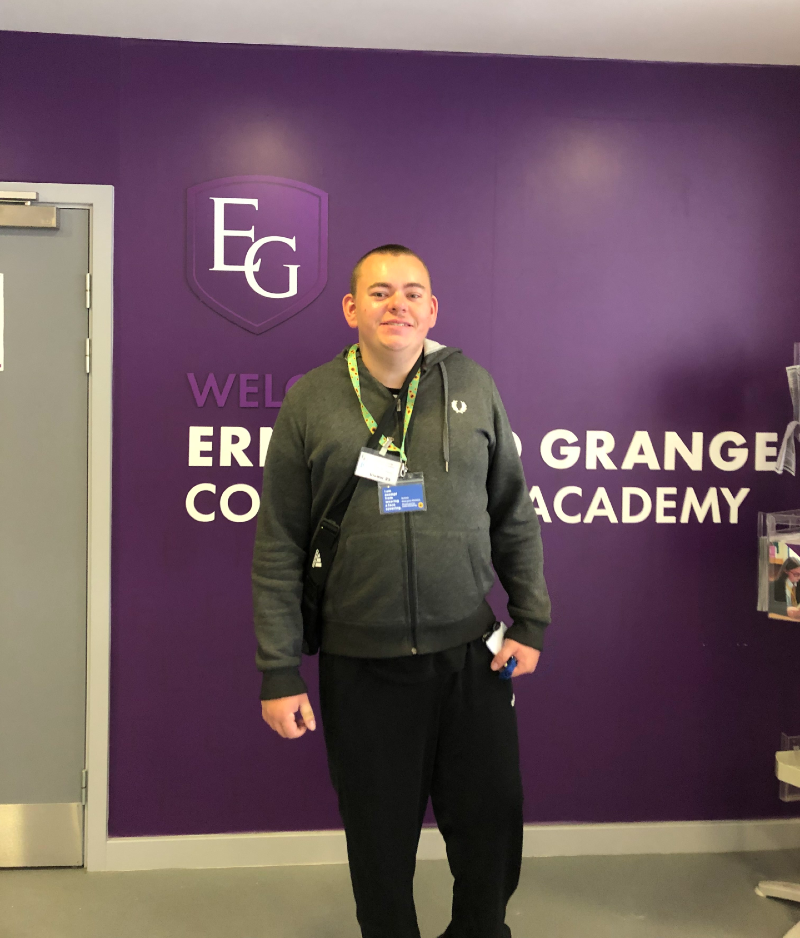 Nathan, a young man with closely cut brown hair wears two lanyards and a crossbody bag as he stands in front of a purple Ernesford Grange sign inside the school. He is smiling on his first day.