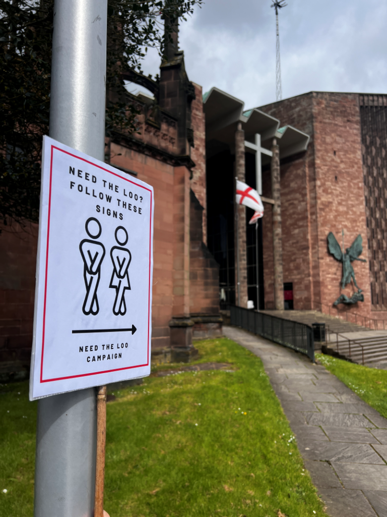 A handmade 'toilets this way' signpost is attached to a lamp post with Coventry Cathedral in the background.