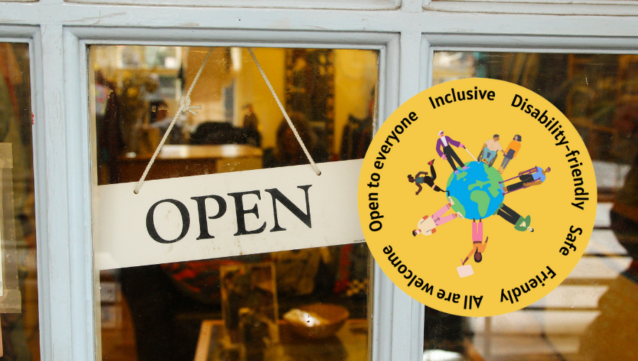 The Inclusive Badge sits in a shop window. It is yellow with an Earth in the centre surrounded by all kinds of people. The text around the edge includes words like Inclusive, Disability-friendly and Open To Everyone.