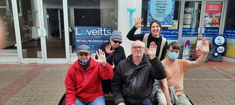 Four people from Nuneaton Speaking Up Group, plus Sophie from Grapevine, smile and wave while they sit on a bench in Nuneaton town centre