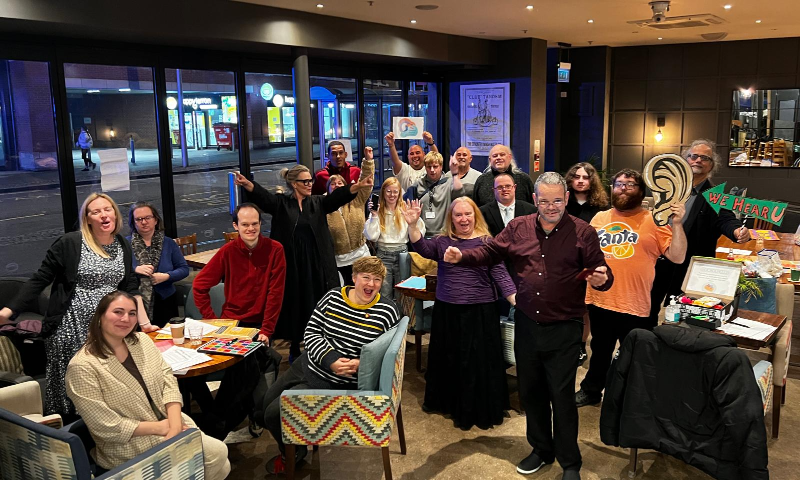 A large group inside a cafe in Coventry city centre smile and cheer at the camera. This is Collaboration Station in action.