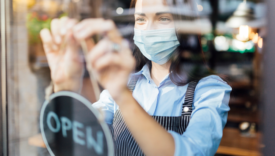 A woman wearing a mask and an apron hangs an Open sign in a shop window