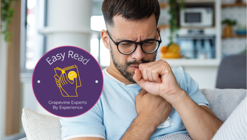 A young white man with dark hair, beard and glasses wears a light blue t-shirt as he sits on his sofa under a blanket coughing into his hand and holding his chest with the other. A purple logo on the image says, "Easy read by Grapevine Experts By Experience."