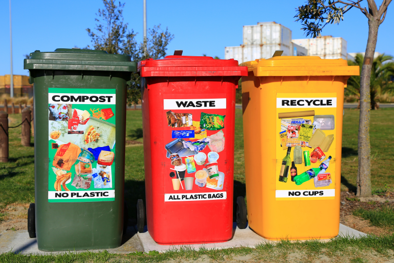 Green, red and yellow litter bins stand in a row in the sunshine on an urban street. They invite people to compost, waste or recycle their rubbish in that order with photos oof what people can put in them on the front of each bin.