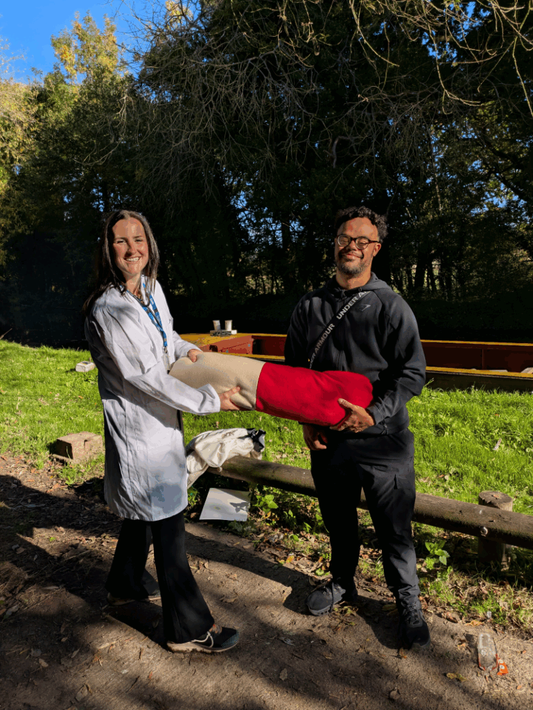 A white woman (Molly) wears a white doctors coat and stethoscope as she smiles and has a tug of war over a giant pain pill with mixed race disabled man Rishard on the canal towpath in the sunshine.