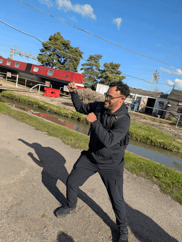 Mixed race disabled man Rishard wears glasses and sports gear as he takes a boxing stance on the canal towpath. He is smiling. 