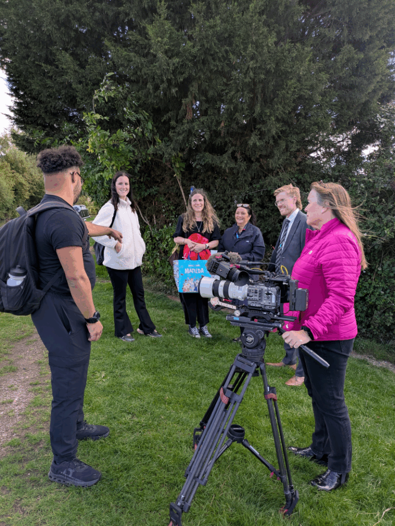 An older white woman in a pink jacket holds a camera on a tripod on a canal towpath and looks on as Rishard chats to Grapevine staff and NHS nurse Petty during a break from interviews.