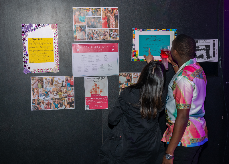 A black couple facing away from the camera hold drinks and read nomination stories stuck to the wall at the venue.