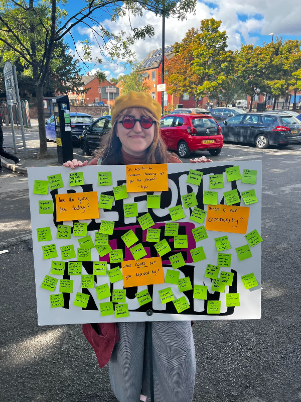 A young women with long hair wears a hat and sunglasses and holds a board covered in post-it note ideas from the community.