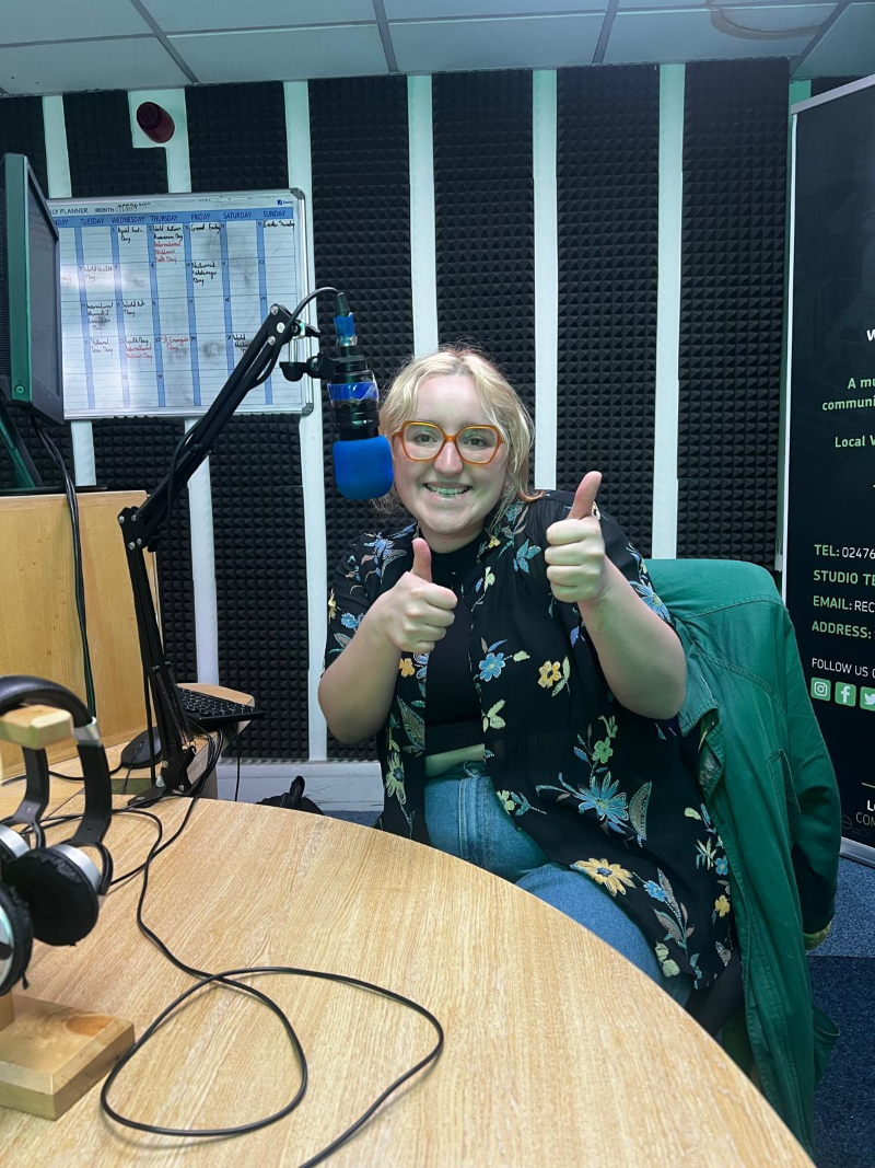 A white woman in her twenties with blonde hair and glasses smiles and gives two thumbs up to the camera while sitting in a radio station studio next to a microphone. She is Sian Lewis from Grapevine.
