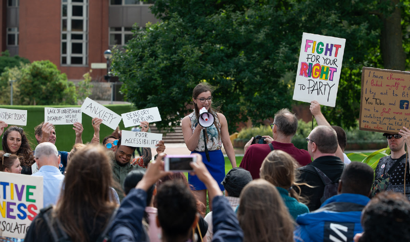 Sophie from Grapevine, a young white woman with long brown hair and glasses, addresses the crowd through a megaphone. People holding placards about isolation are also seen.