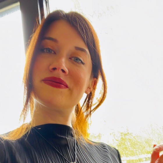 A white woman with dark brown hair wearing red lipstick, a black top and silver chain looks thoughtfully into the camera lens for a selfie against a window during the day.