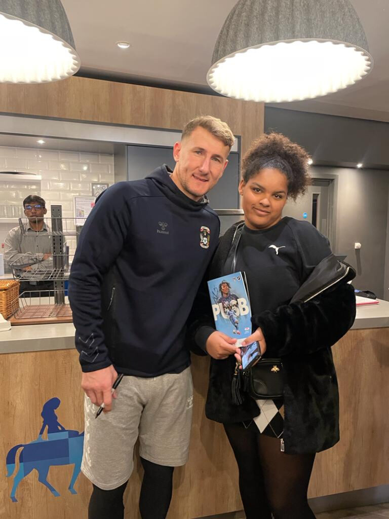 A professional football player wearing a blue Sky Blues hoody poses with a teenage black girl holding the game programme after the match.