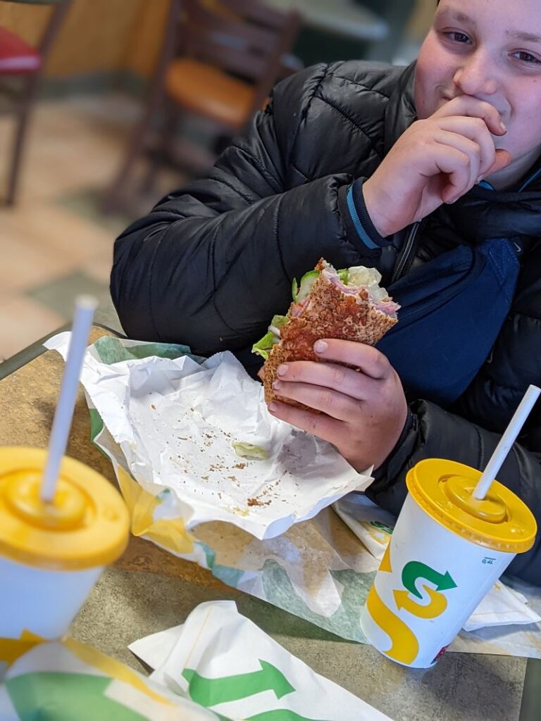 A young teenage boy smiles as he eats a Subway sandwich and has a drink in a local fast food restaurant.