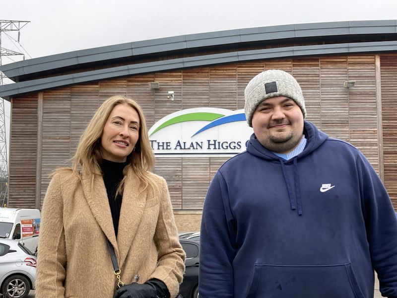 A woman and man wearing warm clothes stand together smiling outside the Alan Higgs leisure centre in Coventry. The building is behind them and the sign can be seen.