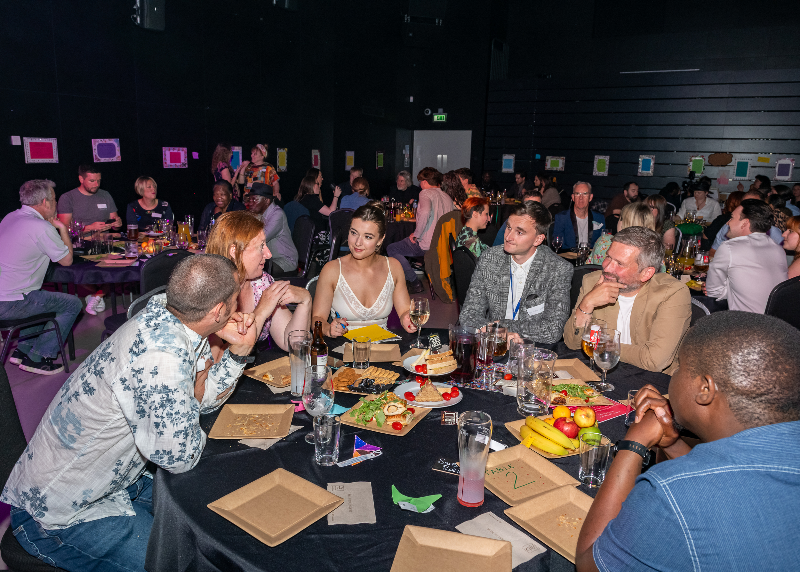 A large event room at night with several tables surrounded by dozens of people in both smart and casual dress, depending how they feel most comfortable.