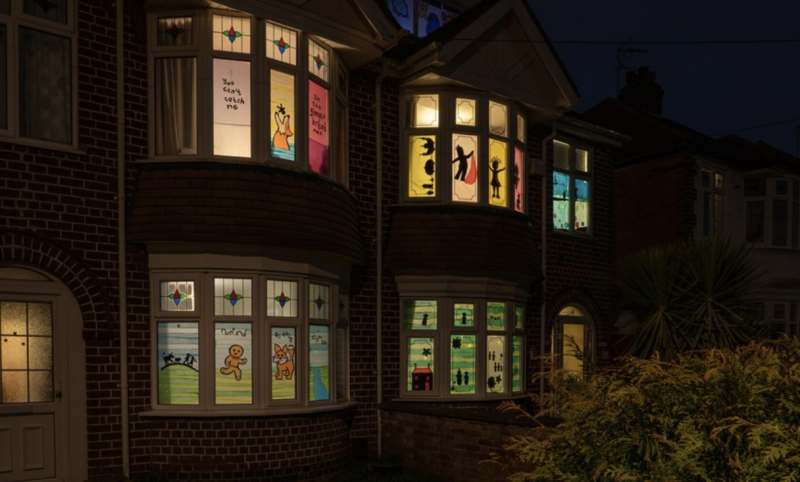 The front of 2 houses in the dark, with bay windows lit up and decorated with colourful images and phrases, including 'you can't catch me', next to a fox coming out of water with a gingerbread man about to fall into the fox's mouth, a gingerbread man running across a green field with blue skies towards a fox cub, silhouettes of houses, stars and cartoon characters.