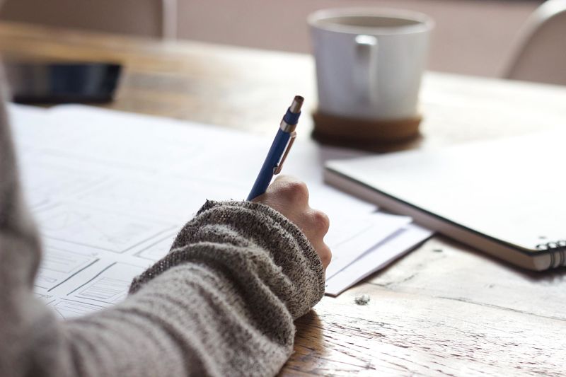 A person writes on paper with a coffee mug on the table