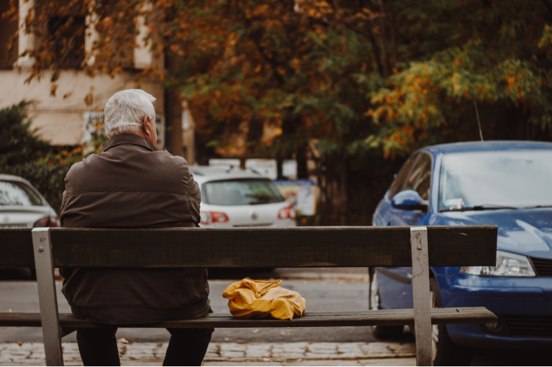 An older man with grey hair sits alone on a bench in an urban street with parked cars in view. Stock image.