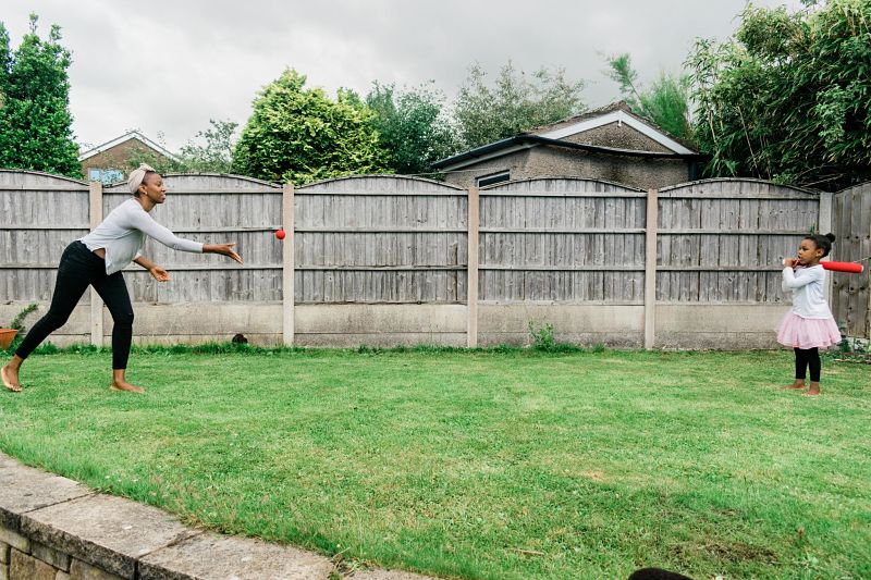 A black woman throws a red ball to a child holding a bat in a back garden on the grass